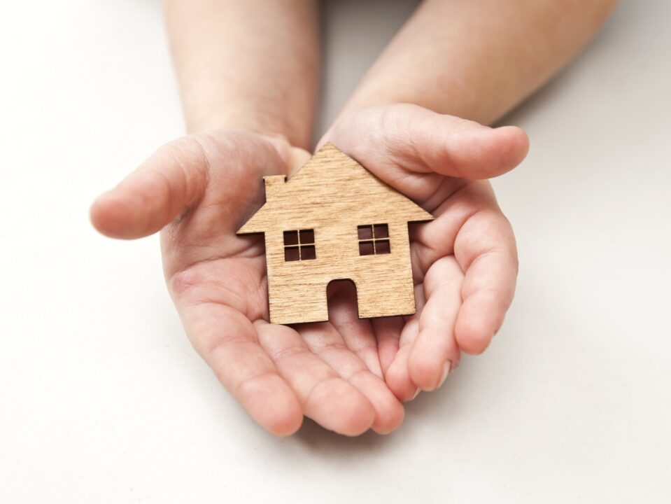 A Little Child Holds Wooden Flat House In His Hands On The White