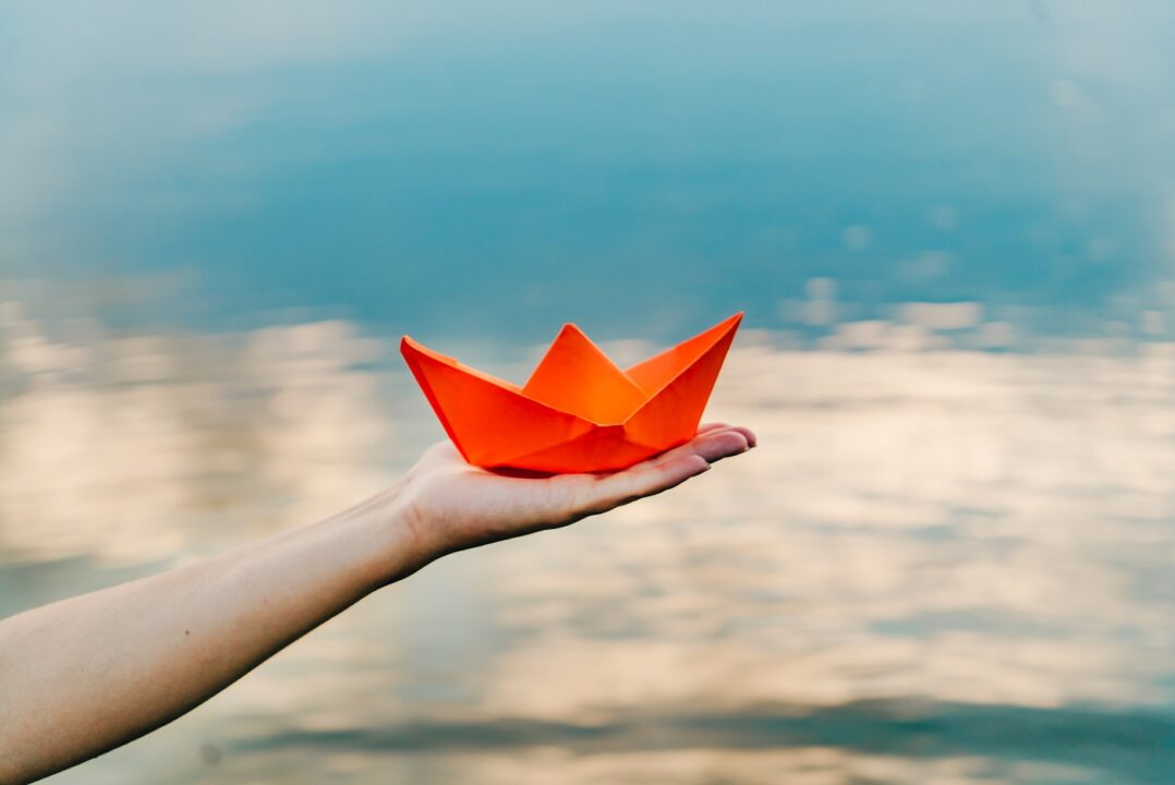 Nice Red Paper Boat On The Hand Of A Young Woman Above The Water In The River Background. Woman Holding Origami Boat Near River. Close Up