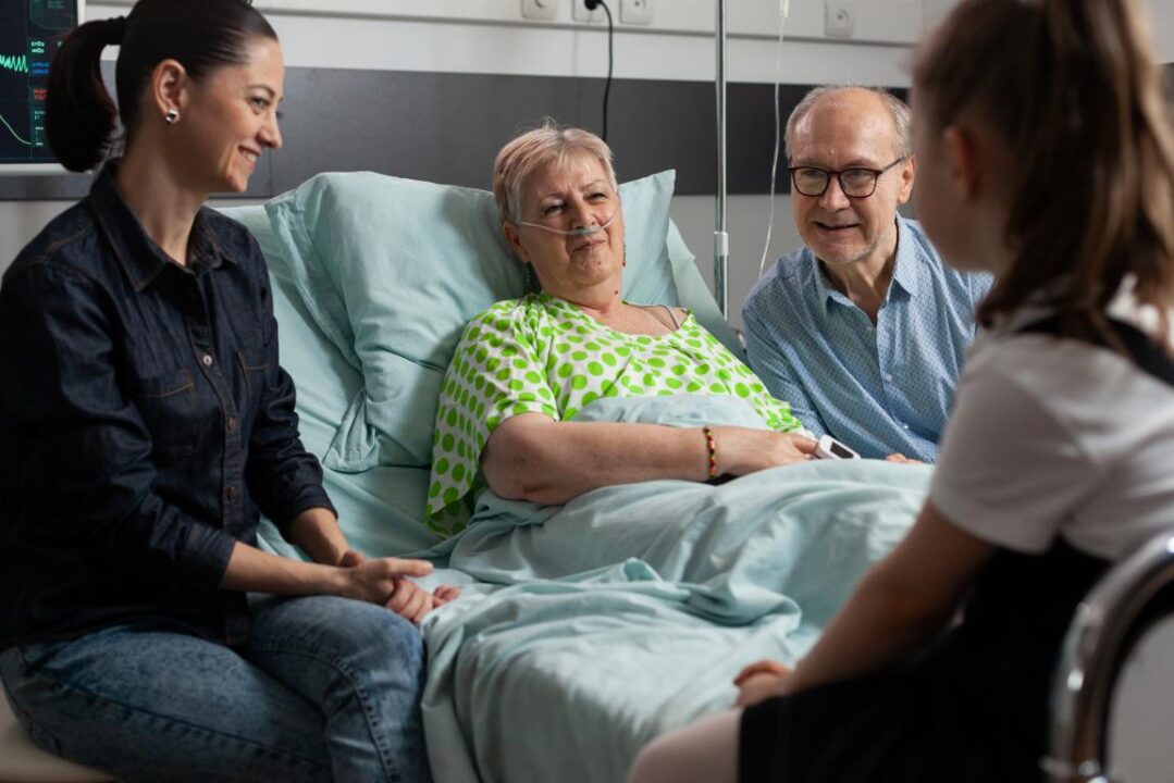 Cheerful Family Supporting Sick Grandmother Visiting Her In Hospital Ward