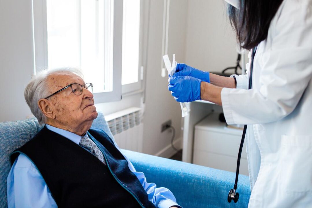 Elderly Man In Blue Shirt And Tie Looking At A Nurse While Opening A Covid Test Home Care