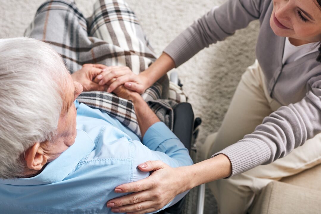 Young Supportive Female Caregiver Sitting By Senior Man In Wheelchair