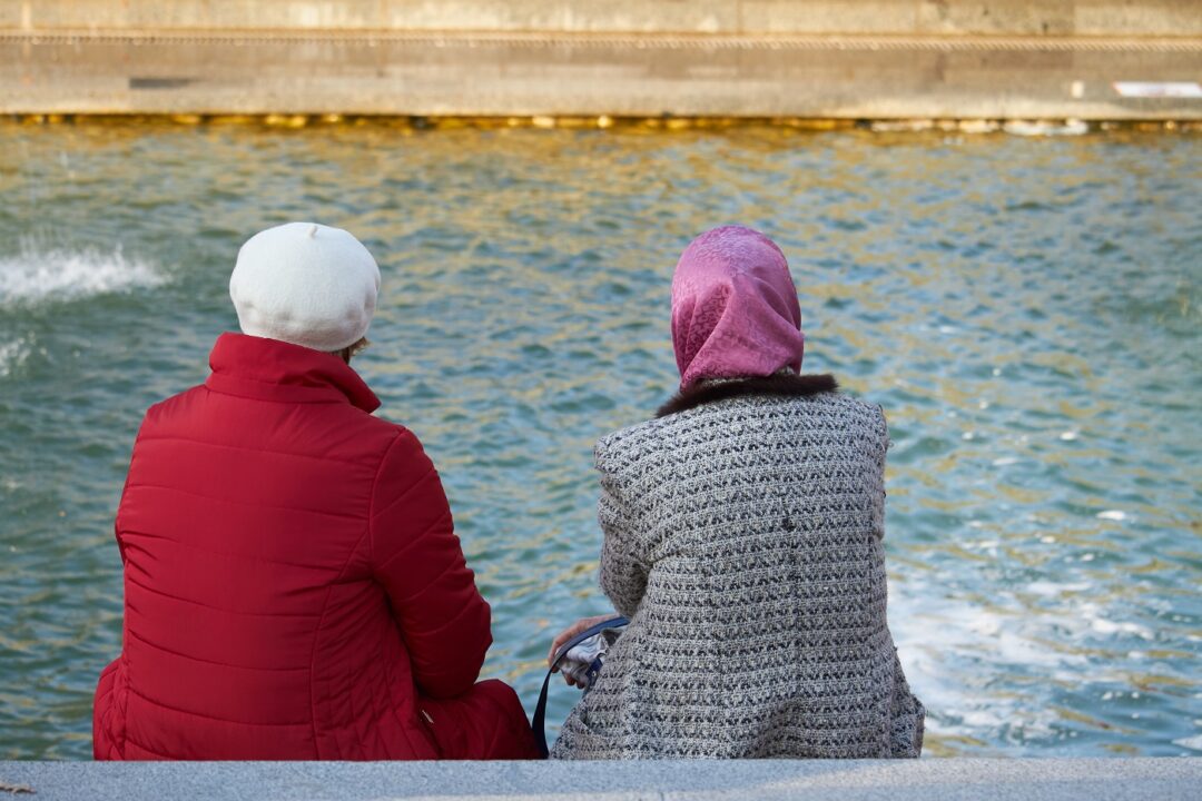 Rear View Of Two Old Women Sitting On A Bench By The Canal In A City Park