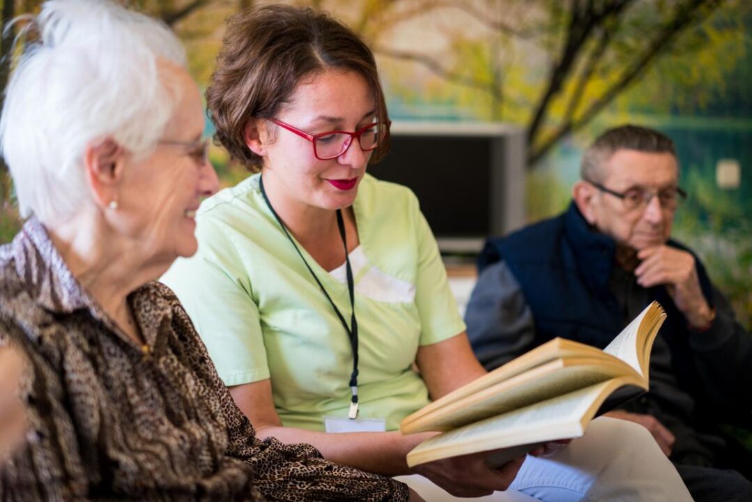 Nurse Reading Book To Elderly Group Of People